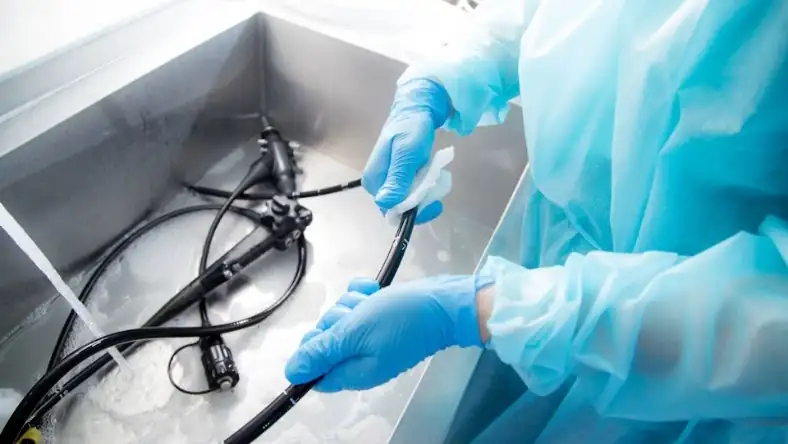 Blue-gloved clinician in a surgical gown cleaning flexible endoscopes in a stainless steel reprocessing basin