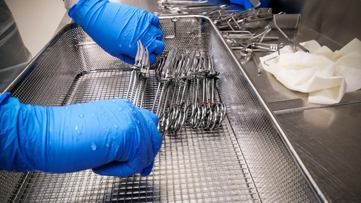 Gloved hands arranging stainless steel instruments in a perforated metal tray in a clinical sterile setting