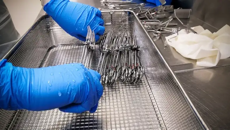 Gloved hands arranging stainless steel instruments in a perforated metal tray in a clinical sterile setting